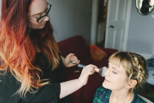 Red-haired woman with glasses applies makeup to a child in a warmly lit home setting