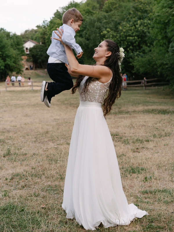 Bride lifting and holding young boy joyfully wearing white gown at outdoor garden wedding venue.