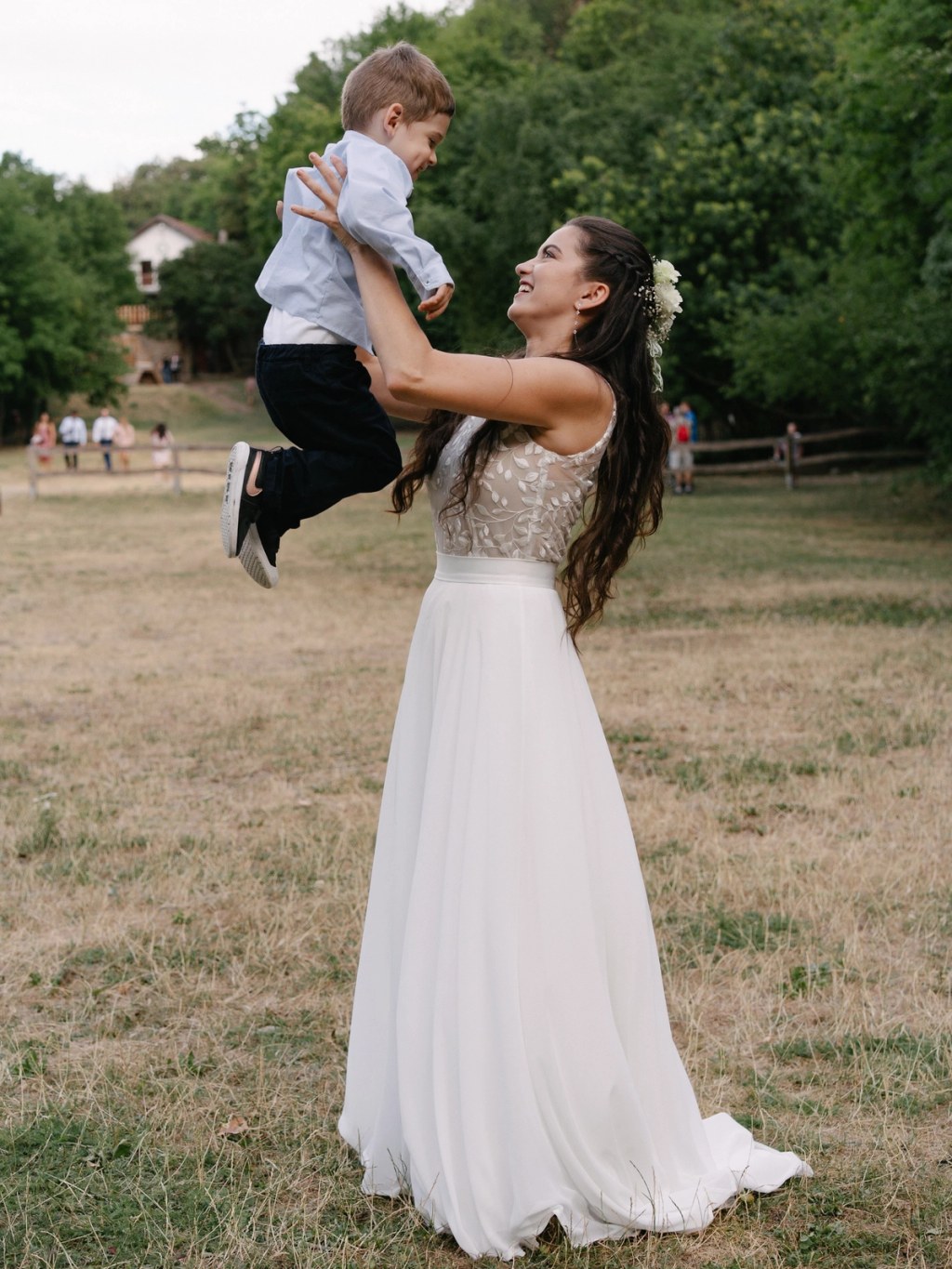 Bride lifting and holding young boy joyfully wearing white gown at outdoor garden wedding venue.