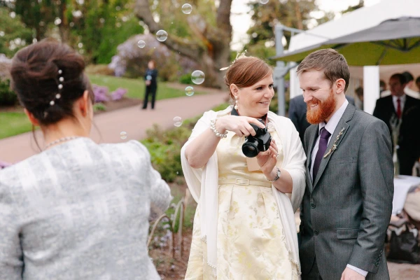 A female wedding guest with a camera laughs alongside a bearded man with a red tie amid floating soap bubbles in the Royal Botanic Garden Edinburgh. Candid documentary wedding photograph.