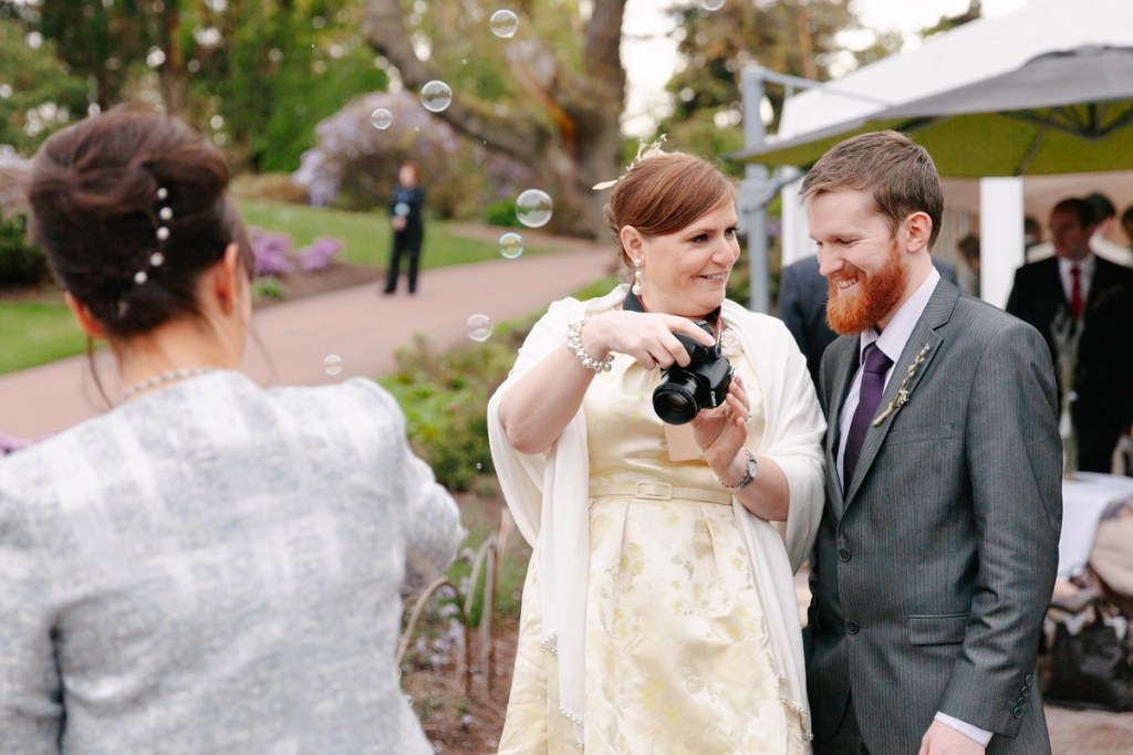 A female wedding guest with a camera laughs alongside a bearded man with a red tie amid floating soap bubbles in the Royal Botanic Garden Edinburgh. Candid documentary wedding photograph.