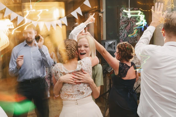 Bride in white lace dances with arms raised in the ballroom, friends and family partying around