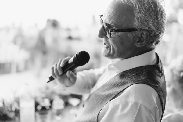 Older gentleman in glasses and tie speaks without a microphone at the celebration — guests slightly blurred