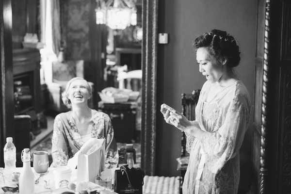 Two women laughing in front of a mirror — lace dress, cosmetics on the table. Black-and-white.