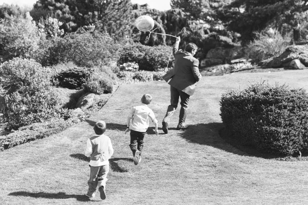 Black and white candid shot of three children running across a lawn at the Royal Botanic Garden Edinburgh, one holding a balloon on a stick. Carefree documentary moment.