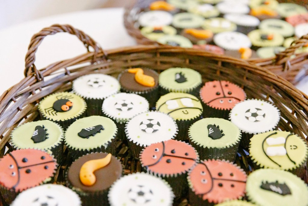 Two wicker trays of decorated wedding cupcakes with colourful fondant toppers including football, ladybird, and animal motifs. Personalised wedding cake detail.