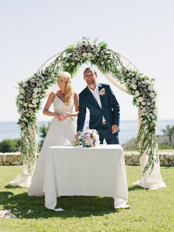 Bride and groom smiling under the white floral gate — signing table with bouquet and sea view