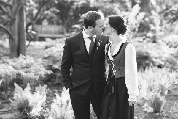 Black and white image of the couple kissing amid lush garden ferns and shrubs, the bride in her Norwegian bunad. Romantic documentary wedding portrait at the Royal Botanic Garden Edinburgh.