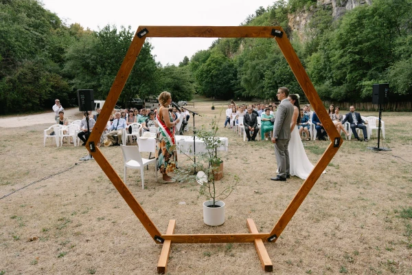 Outdoor garden wedding ceremony with wooden hexagon arch and daisy flower arrangement under trees.