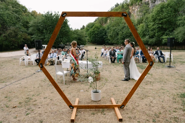 Outdoor garden wedding ceremony with wooden hexagon arch and daisy flower arrangement under trees.