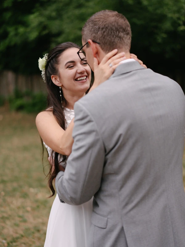 Bride smiling joyfully while groom holds her at outdoor wedding with green background and trees.