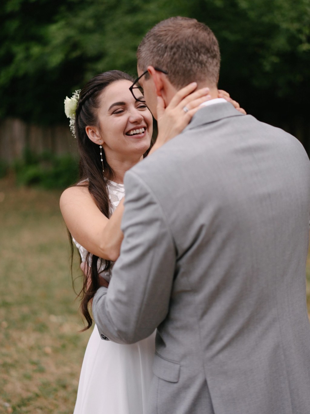 Bride smiling joyfully while groom holds her at outdoor wedding with green background and trees.