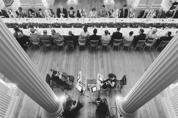 Elegant wedding dinner from above — guests seated, orchestra playing in a classical institutional hall
