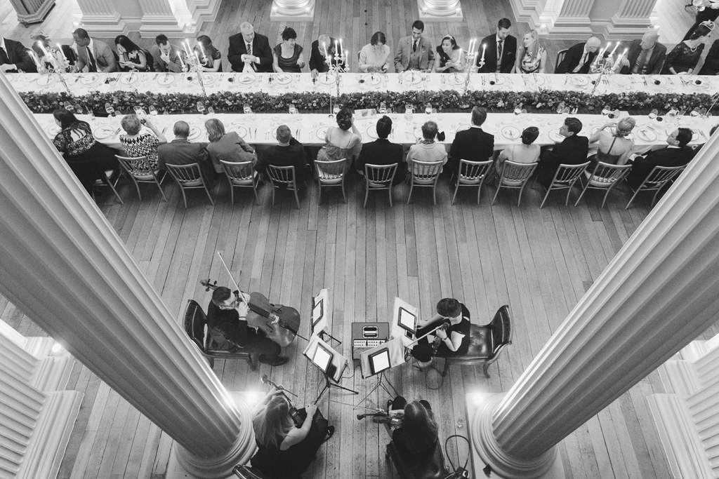 Elegant wedding dinner from above — guests seated, orchestra playing in a classical institutional hall