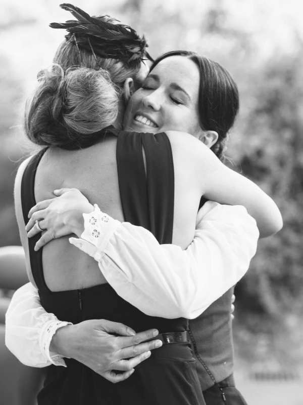 Black and white close-up of the bride smiling with closed eyes as she shares a warm embrace with a guest wearing a feathered fascinator at the wedding. Documentary wedding photography.
