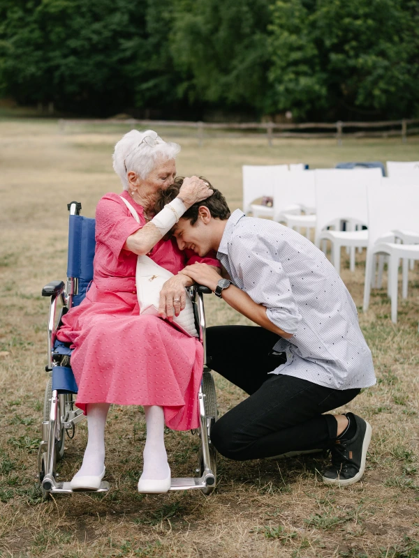 Candid photo of an elderly woman in a pink dress kissing the forehead of her grandson at an outdoor wedding reception.