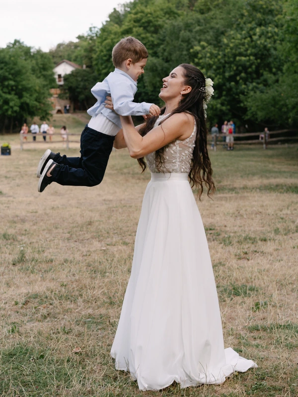Bride playfully holding small boy in air with white wedding dress and family in background.