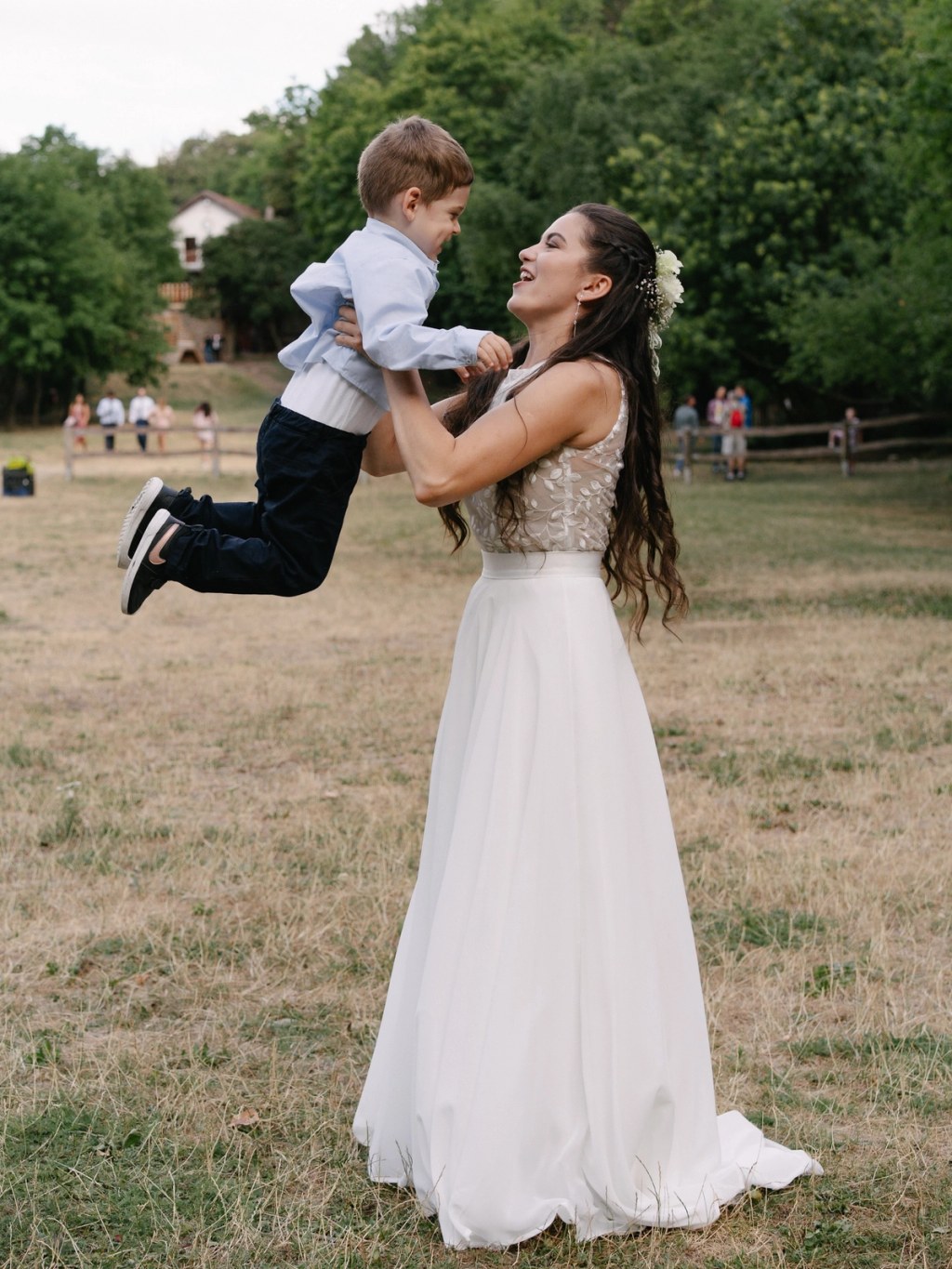 Bride playfully holding small boy in air with white wedding dress and family in background.