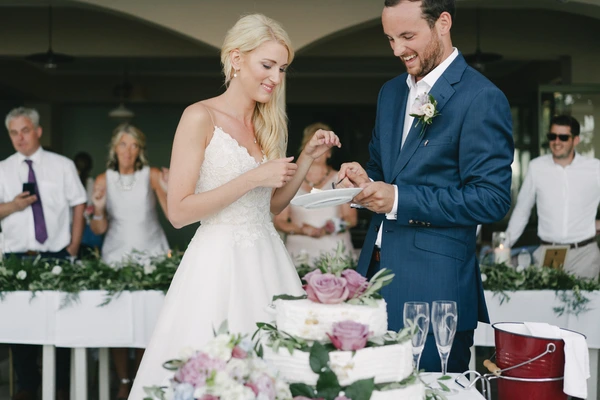 Bride and groom smiling at the wedding cake at the reception — scattered guests, floral table, warm light