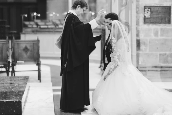 Priest blesses the veiled bride in the church — groom beside her. Black-and-white ceremony.