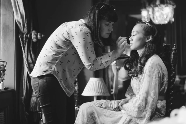 Makeup artist carefully prepares the seated bride's face in warm lamp light at the vanity table