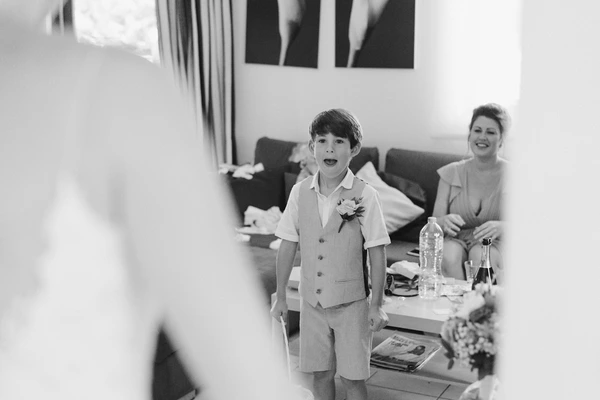 Boy in wedding vest and tie stands smiling in the room, woman smiling on the sofa beside him