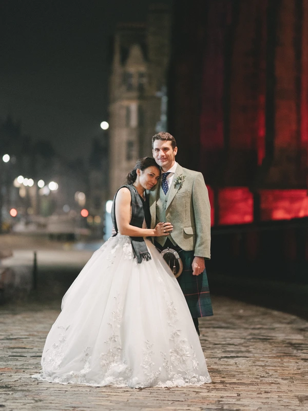 Couple smiling at each other in a night city square — groom in tartan, bride in white lace