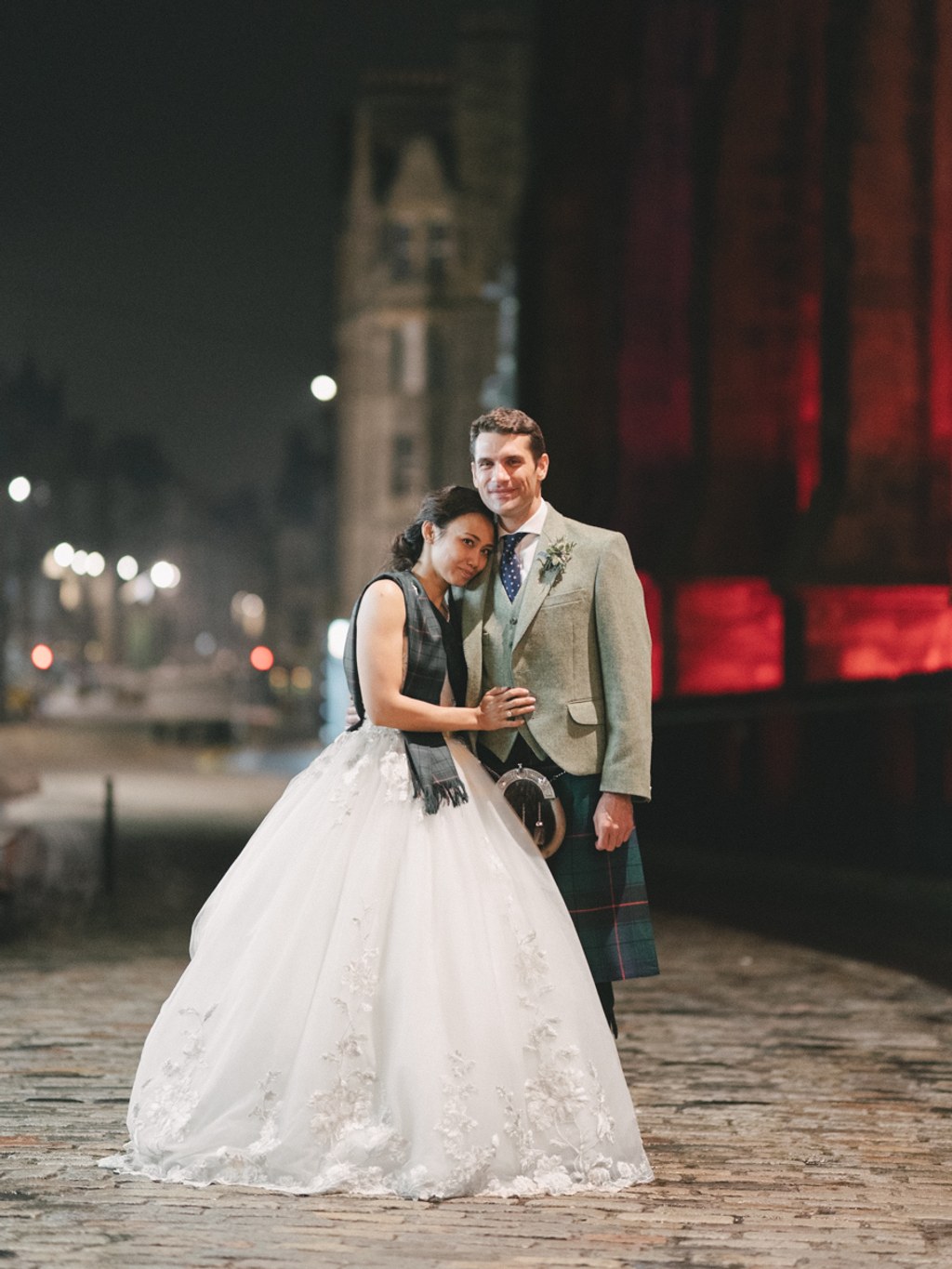 Couple smiling at each other in a night city square — groom in tartan, bride in white lace