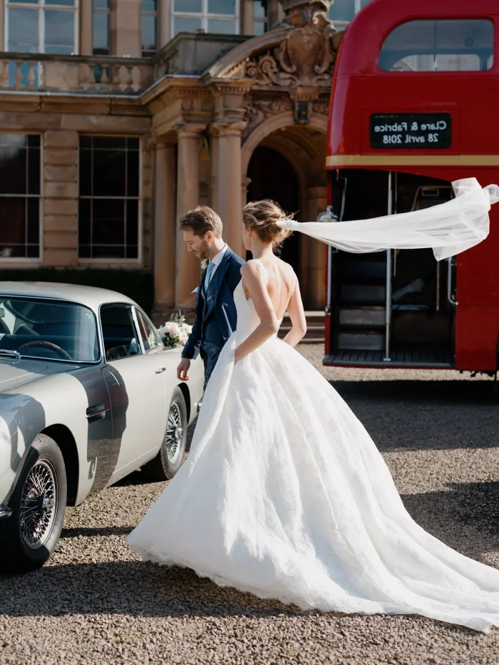 Bride and groom walk past vintage Aston Martin at grand estate, veil billowing in wind, red double-decker bus behind them.
