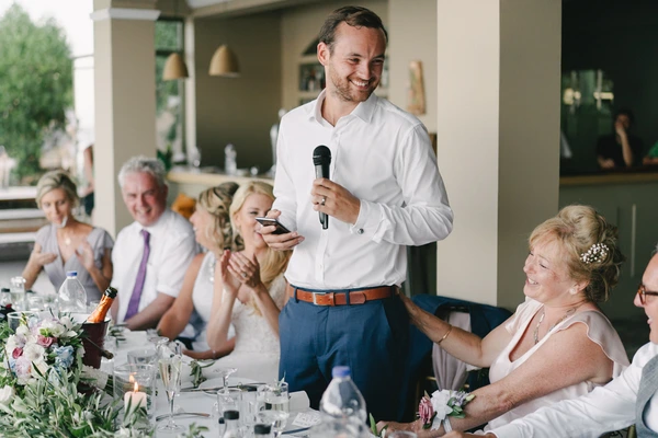Groom at the microphone giving his speech at the wedding dinner — white shirt, blue trousers, guests laughing