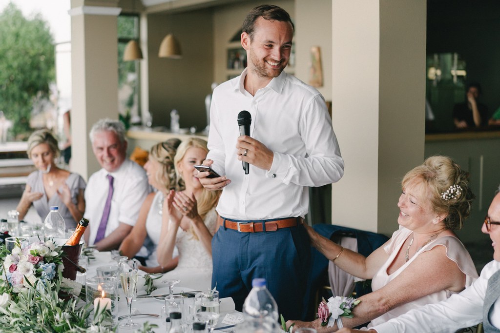 Groom at the microphone giving his speech at the wedding dinner — white shirt, blue trousers, guests laughing