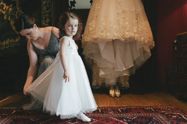 Woman adjusts a little girl's white tulle dress in an ornate dark salon, bride visible behind