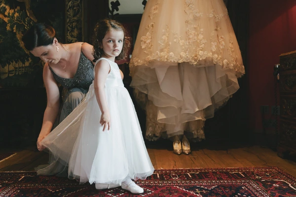 Woman adjusts a little girl's white tulle dress in an ornate dark salon, bride visible behind