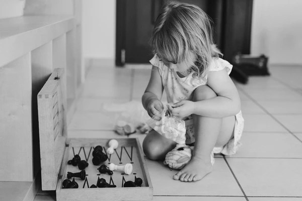 Little girl plays with chess pieces on the floor in a white dress, concentrated gaze downward
