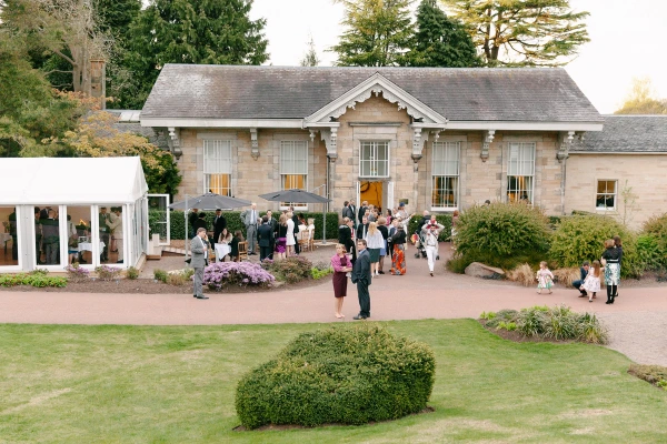 Aerial-style view of wedding guests mingling outside the stone venue building at the Royal Botanic Garden Edinburgh in the late afternoon light, with a marquee visible to the side. Documentary wedding photograph.