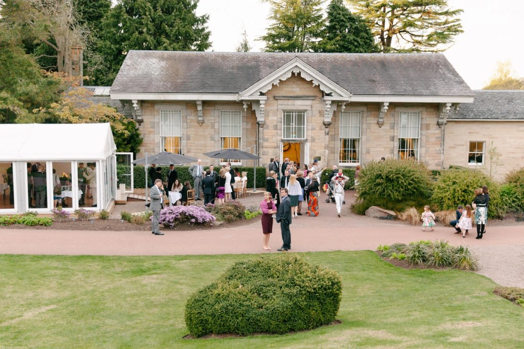 Aerial-style view of wedding guests mingling outside the stone venue building at the Royal Botanic Garden Edinburgh in the late afternoon light, with a marquee visible to the side. Documentary wedding photograph.
