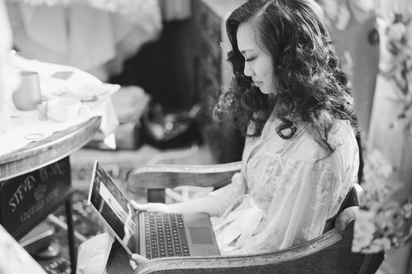 Bride at a laptop during wedding preparations, white lace dress, pensive look at the screen