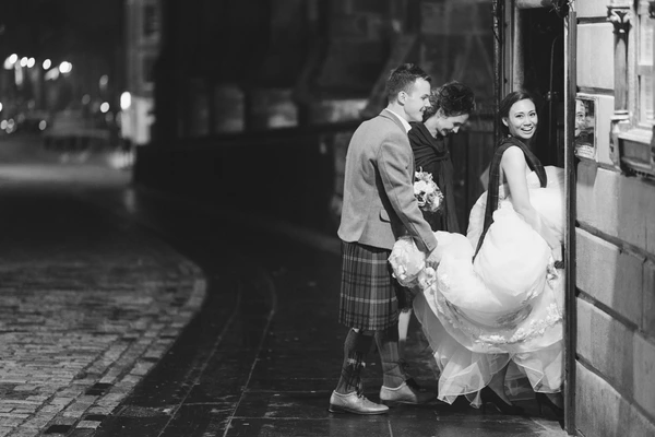 Groom and bride hand in hand before a doorway at night — city lights and urban setting