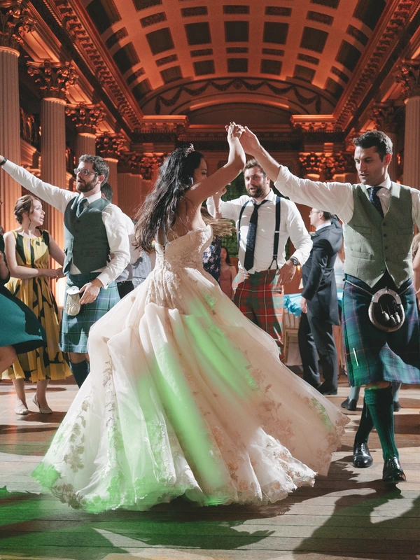 Bride spins her white lace gown dancing — guests in tartan and formal wear, orange-lit ballroom