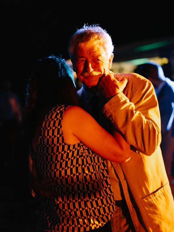 Elderly man dancing joyfully at wedding celebration — warm golden light, emotional moment, patterned shirt.