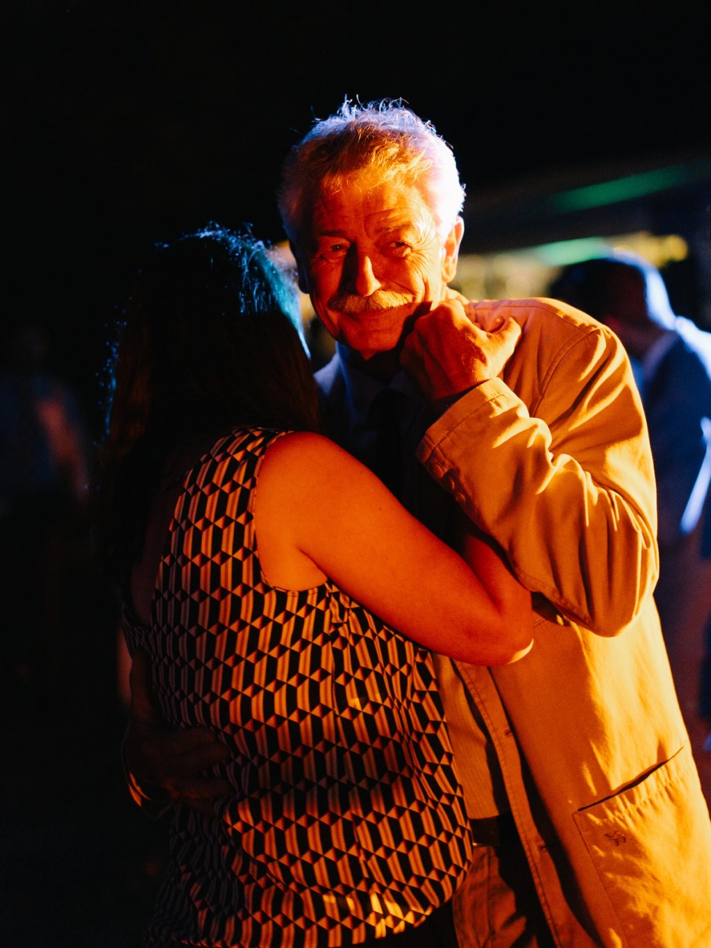 Elderly man dancing joyfully at wedding celebration — warm golden light, emotional moment, patterned shirt.