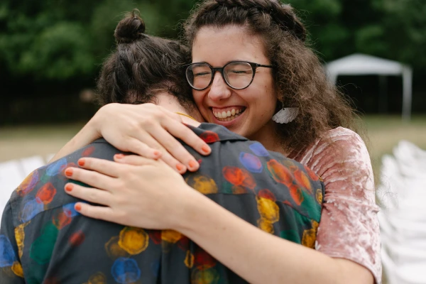Woman with glasses hugging guest in patterned shirt at outdoor wedding celebration with joyful expression.