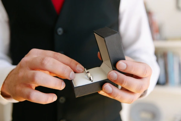 Groom in a dark waistcoat and red tie holding open a ring box with two wedding rings inside. Close-up pre-ceremony detail.
