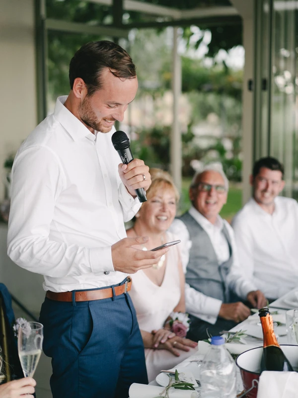 Groom smiles giving his speech to family and friends at the table — green garden backdrop