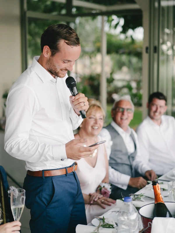 Groom smiles giving his speech to family and friends at the table — green garden backdrop