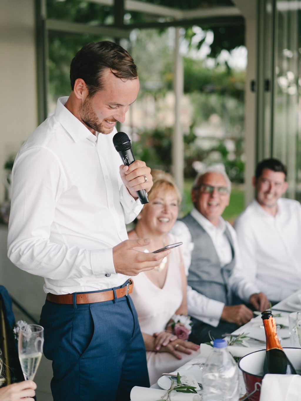 Groom smiles giving his speech to family and friends at the table — green garden backdrop