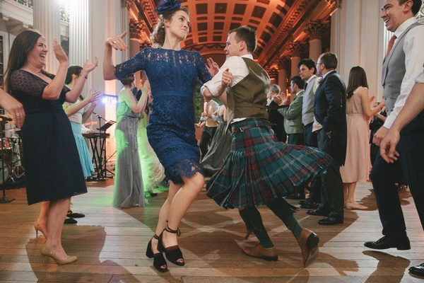 Joyful dance in an elegant columned hall — woman in blue lace and man in tartan kilt at centre