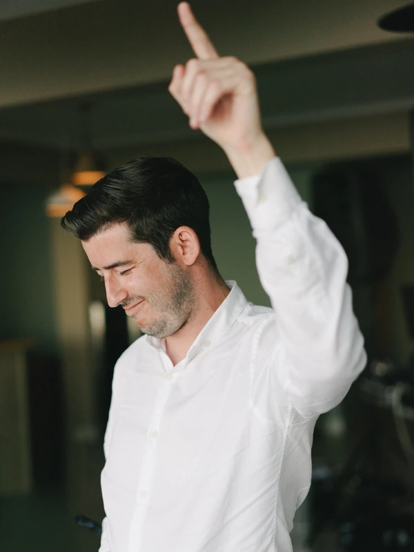 Groom in white shirt with a wide grin mid-gesture — celebratory moment during the wedding preparations