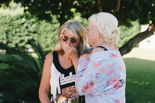 Two women before a green garden backdrop — one in sunglasses, moved expression; one in floral dress
