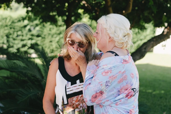 Two women before a green garden backdrop — one in sunglasses, moved expression; one in floral dress